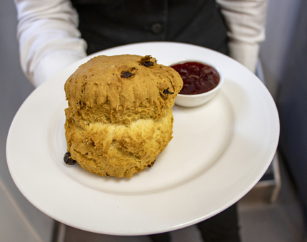 A Waiter holding a plate containing a large fruit scone and a small dish of jam. 