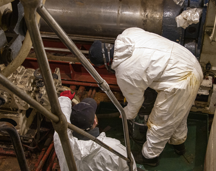 Two members of the Maintenance team vacuuming in the Stabiliser Compartment, which contains large machinery. 