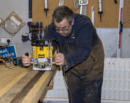 A man in the Maintenance team routering a handrail with machinery on a workbench.