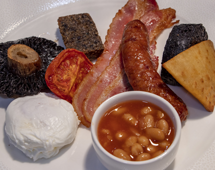 A plate of a full Scottish cooked breakfast with poached egg, black pudding, tomato, bacon, sausage, hash brown, haggis and mushroom.