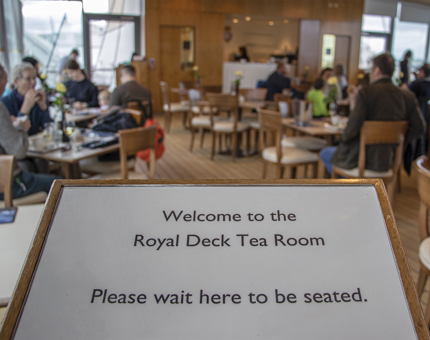 A board reads 'Welcome to the Royal Deck Tea Room. Please wait here to be seated'. In the background, groups of visitors are seated at tables enjoying tea. 