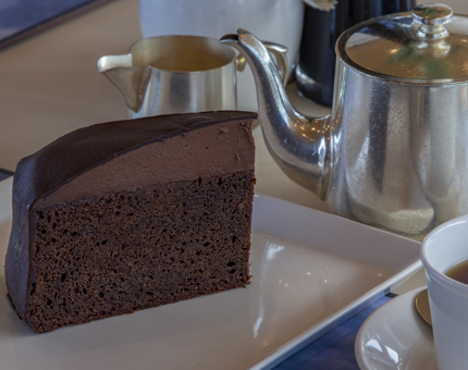 A large slice of chocolate fudge cake sitting on a plate, next to a teapot, jug of milk and cup and saucer. 