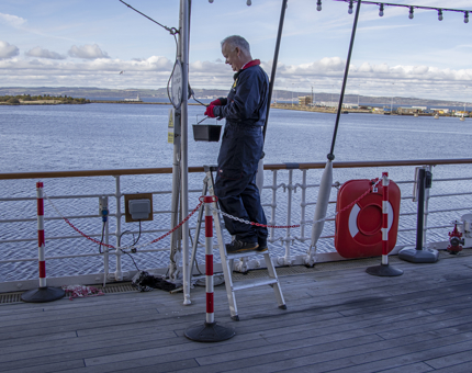 A man standing on a ladder holding a rollerbrush and paint bucket as he paints a support on the Verandah Deck.