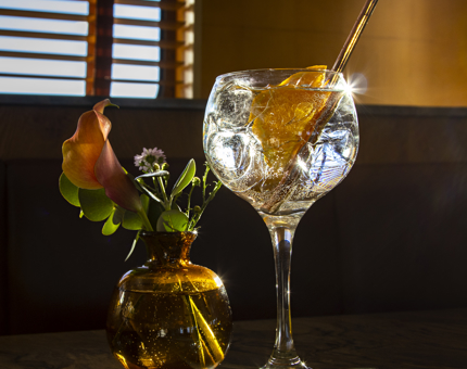 A small round vase of flowers sitting next to a large gin and tonic. A window is in the background. 