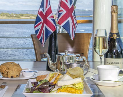 The Coronation Cream Tea served in the Royal Deck Tea Room. A scone, Coronation chicken sandwich and salad, pot of tea and bottle of Gusbourne wine is sitting on the table. Union Jack flags are in a vase on the table.