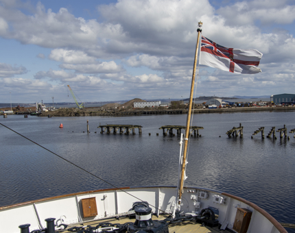 The view from the stern of Britannia. The flag is blowing in the wind and in the background, in the Firth, stands an Antony Gormley figure statue '6 Times'. 