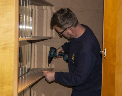 A member of the Maintenance team holding a drill to fix shelving in a cupboard. 