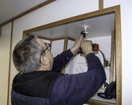 A man in the Maintenance team holding a drill to fix a porthole deadlight.