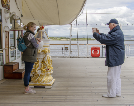 A Yottie takes the photograph of a visitor standing next to the ornate gold painted compass binnacle. 