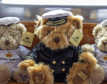A plush Captain teddy bear wearing a Captain's uniform sitting between two soft Yottie teddy bears, wearing white Yottie outfits. 