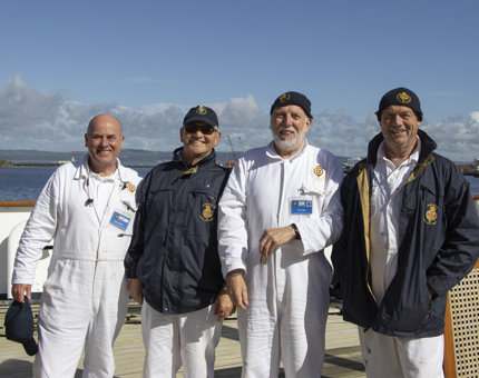 A group of four smiling Yotties standing outside on deck against a blue sky.