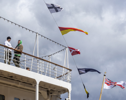 Multi-coloured dress flags are fluttering in the breeze, next to the Verandah Deck where two visitors lean on the handrail admiring the view. 