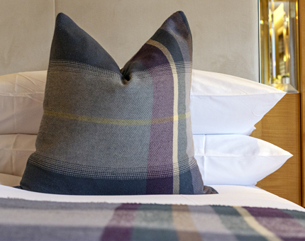 A blue and purple tartan cushion sitting on a bed inside a Fingal cabin. A matching runner and pillows are also on the bed. 