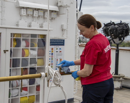 A Housekeeper polishing a brass pole next to the Flag Box on the Bridge.