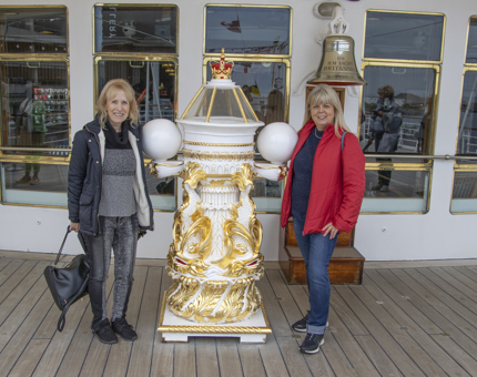 Two women visitors stand smiling either side of Britannia's compass binnacle. 