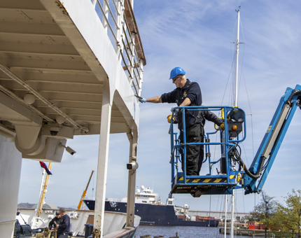Two men on a cherry picker working on the outside of the hull. 
