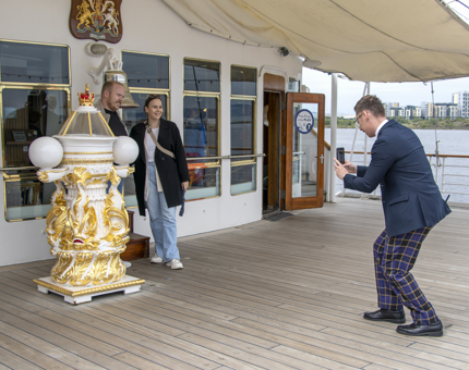 Visitor Assistant taking a photograph of a couple standing next to Britannia's binnacle.