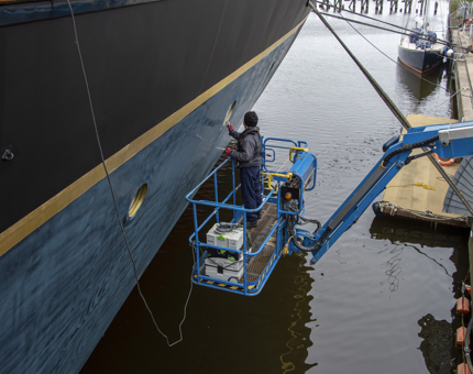 A member of the Maintenance Team is on a cherry picker working on applying filler to an outside porthole.