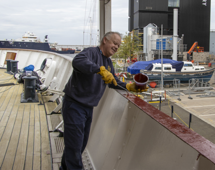 A man painting the top of the ship's side with a red-brown paint.