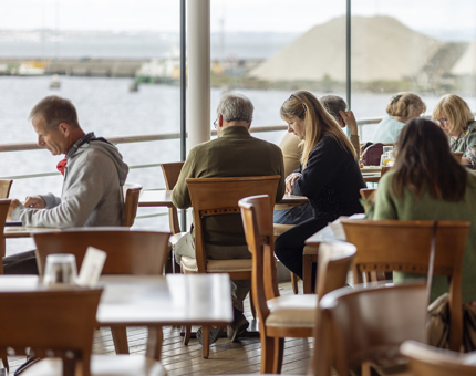 Visitors sitting at tables in the Royal Deck Tearoom. There is a view of the Port of Leith in the background. 