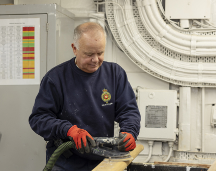 A maintenance team member holding an electric sander to sand down a wooden handrail.