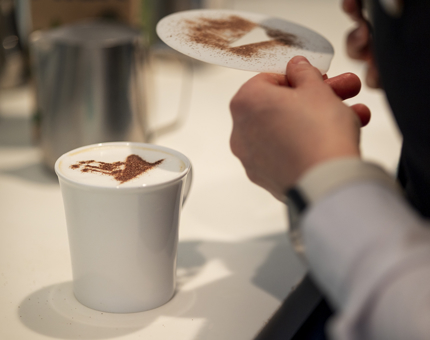 In the Tearoom, a cappuccino is being prepared, a chocolate stencil of a ship in being put on the top of it. 