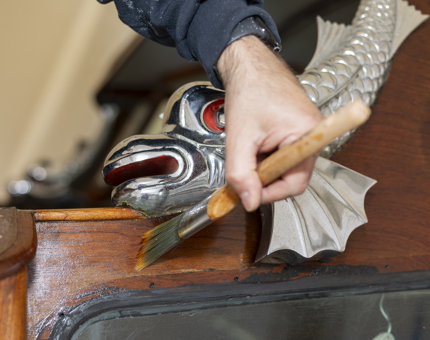 A close up of a hand holding a paint brush, varnishing the Royal Barge.