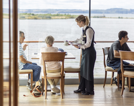 In the Royal Deck Tearoom, there is a server placing a teacup onto a table for two visitors. There is a view of the Port of Leith in the background. 