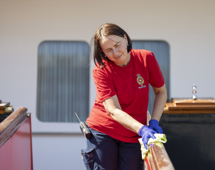 A Housekeeper wearing a red shirt and blue gloves, holding a yellow cloth, wiping down the handrail of the Royal Brow.
