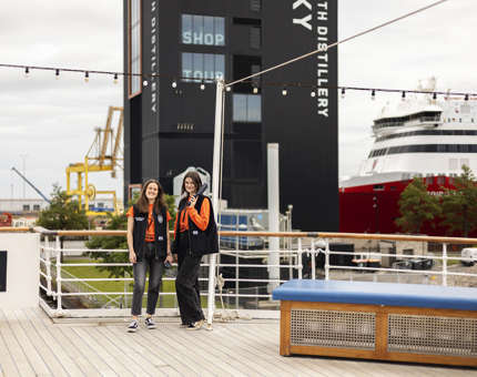Two women listening to the audio guide handsets on the Verandah Deck. The Port of Leith Distillery is in the background. It is a tall black building. 