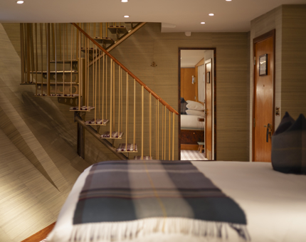 A double bed with white sheets and a tartan blanket. There is a staircase in the background.