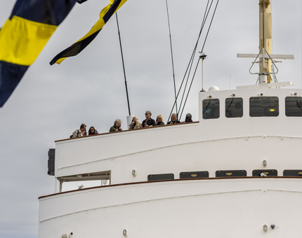 A view of the Bridge from below. There are people listening to their audio guide handsets. There are flags flying from the mast in the foreground. 
