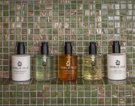 Five bottles of Noble Isle bath and body products on a green tiled shelf.