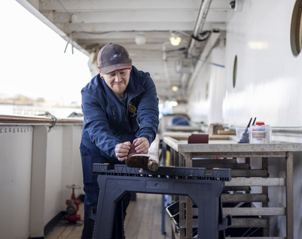 On deck, a member of the Maintenance team is sanding a handrail.