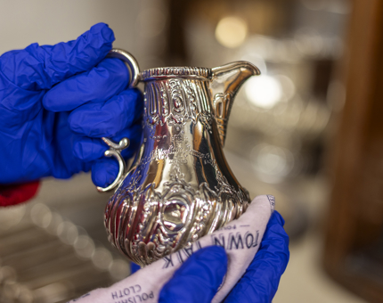 Hands wearing blue gloves holding a small silver jug and polishing it with a pink cloth. 