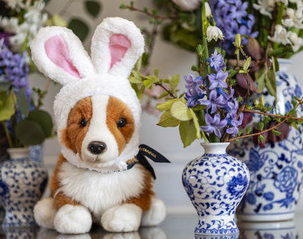 A corgi plush toy with whit bunny ears on its head. There a blue and white vases with purple and white flowers next to it. 