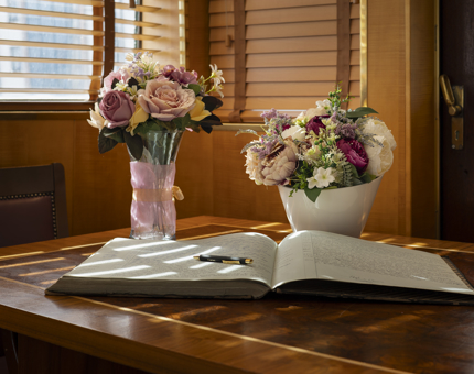 An open log book on a wooden table. There are two vases in the background with pink and white flowers in them.