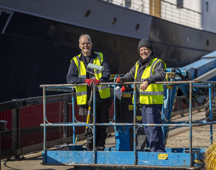 Two men on a blue cherry picker wearing high-vis vests. one man is holding a paint roller. There is a ship in the background.