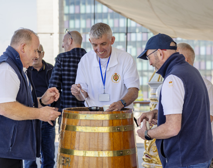 Three Ex Royal Yachtsmen gathered around a rum tub pouring it into small cups. 