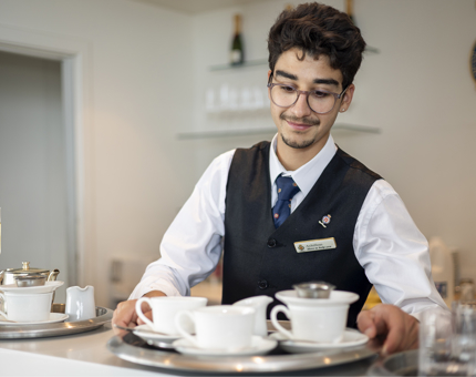 A Hospitality Server is behind the bar in the Royal Deck Tearoom in the Port of Leith. He is picking up a tray with cups and saucers.
