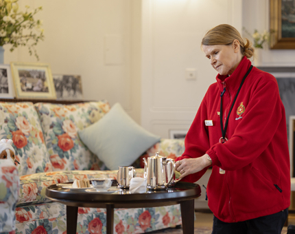 A Housekeeper wearing a red jacket is polishing a silver teaset on a small table in the State Drawing Room. 