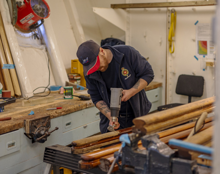 A Maintenance man leaning over wooden handrails, he is removing varnish with a heat gun and scraper.