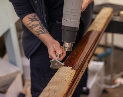 Hands holding a heat gun and scraper, removing varnish from a wooden handrail.