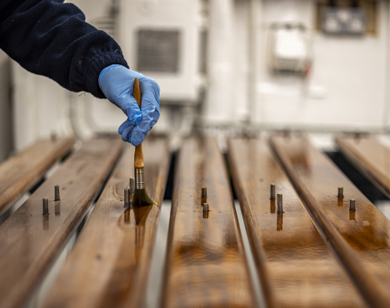 A hand with a blue glove on, holding a paint brush to varnish handrails. 