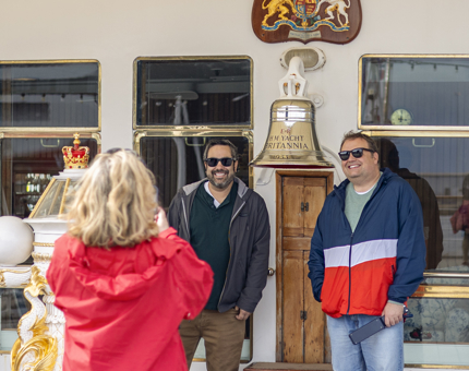 A Visitor Assistant wearing a red jacket taking a photo of two visitors who are standing next to Britannia's Bell. 