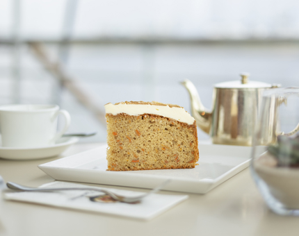 A slice of carrot cake on a table with a pot of tea and teacup. 