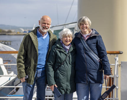 A group of three visitors pose for a photo on the Bridge. The ship's funnel is in the background and the sun is shining. 