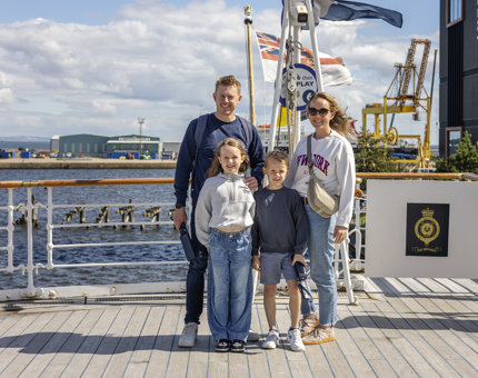 A family posing for a photo on The Royal Yacht Britannia's Verandah Deck in Edinburgh. The Port of Leith is in the background. 
