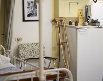 Inside the onboard Sick Bay. There is a hospital bed, a floral print chair, a wooden crutch, a fridge an eye test chart and bottles of medicine in the room. 