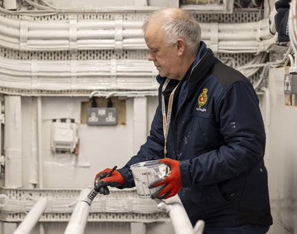 A man wearing red gloves holding a pot of white paint and a paint brush painting poles white. 